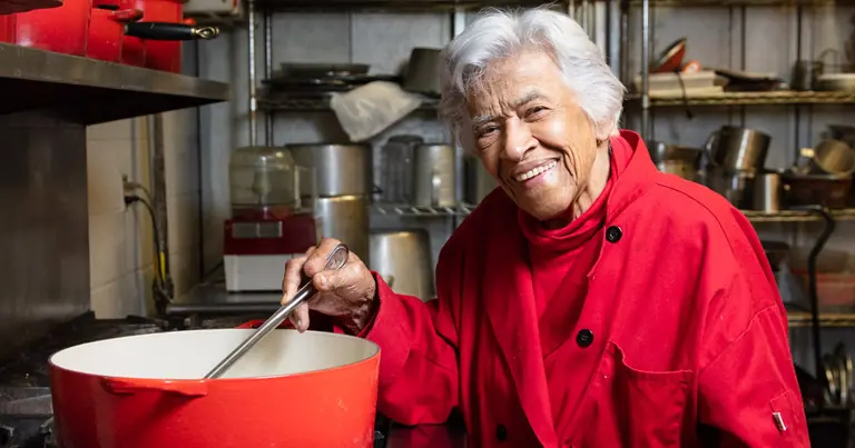Chef Leah Chase in Dooky Chase's kitchen, in a red chef's coat, stirring a large red pot.