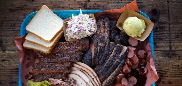 A Franklin Barbecue tray: sliced brisket, sausage and ribs on butcher paper with white bread, pickles and onion.