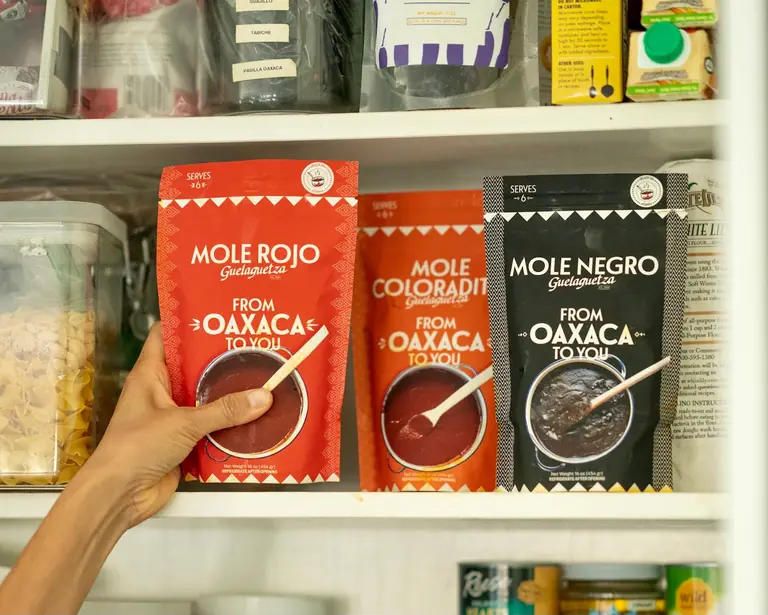 Packets of Guelaguetza mole — rojo, coloradito and negro — on a shop shelf, with a hand reaching for one.