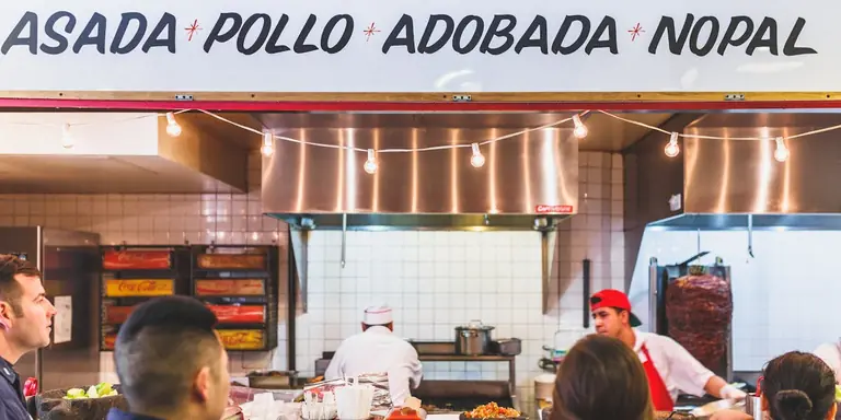 The counter at Los Tacos No.1: staff working the griddle beneath a menu board reading ASADA, POLLO, ADOBADA, NOPAL.