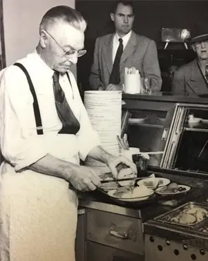 An archival black-and-white photograph of a cook in shirtsleeves and braces working the counter at Philippe the Original.